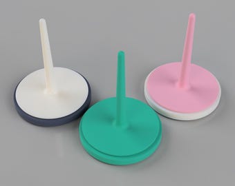 May include: Three colorful, round, ceramic pin holders. One is teal, one is pink, and one is white with a navy blue base. Each has a central, vertical, cylindrical pin. The pin holders are on a gray surface.