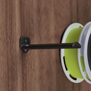 May include: A black metal wall-mounted spool holder with a single spool of green filament attached. The holder is mounted on a wood panel.