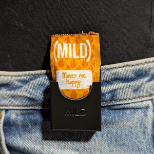 Puede incluir: Un soporte negro con la palabra "MILD" en relieve en la parte delantera, que sostiene un sobre de salsa naranja. El sobre tiene la palabra "MILD" en blanco y la frase "Makes me happy". El soporte está sujeto a unos vaqueros azules.