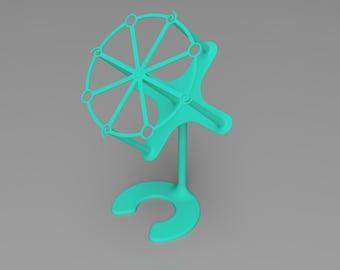 May include: A teal-coloured, 3D-printed object with a circular, wheel-like top and a curved base. The wheel has spokes and small circular openings. The object stands upright on a U-shaped base, connected by a vertical stem.