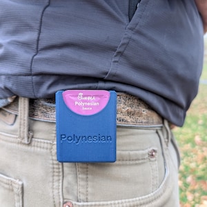 May include: A blue, rectangular holder with the word "Polynesian" embossed on the front. The holder contains a pink packet of Polynesian sauce. The holder is clipped to a belt loop on a pair of khaki pants.