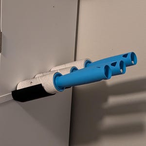May include: A wall-mounted lint roller holder with three blue handles. The holder is attached to a white cabinet and has a black backing. The lint rollers are white with a gray pattern.
