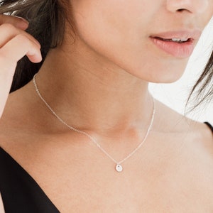 Peut inclure: Un collier en argent délicat avec un petit pendentif rond. Le collier est porté sur un haut noir, créant un contraste qui met en valeur le bijou. La chaîne est fine et le pendentif est simple, ce qui en fait un accessoire polyvalent.