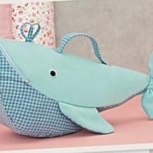 Puede incluir: Un bolso en forma de ballena azul claro con un estampado de cuadros en la cabeza y un cuerpo azul claro liso. Cuenta con un asa, una cremallera y una pequeña cola. El bolso mide aproximadamente 30 cm de largo.