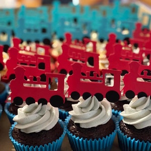 Könnte beinhalten: Nahaufnahme eines Tabletts mit Schokoladen-Cupcakes, die mit weißem Frosting und roten Lokomotiv-Toppern verziert sind. Die Cupcakes befinden sich in blauen Papierförmchen.