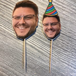 May include: Two face cutouts of a smiling man wearing glasses, one with a party hat. The cutouts are attached to wooden skewers, likely for use as cake toppers or party decorations. The background is a wood-grain surface.