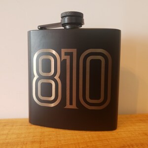 May include: Black flask with the number "810" in a silver font. The flask has a black screw-on cap and a hinged, attached cap cover. The flask is rectangular with rounded corners.