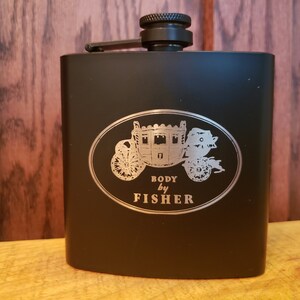 May include: Black flask with a silver oval design featuring a horse-drawn carriage and the words "BODY by FISHER". The flask has a black screw-on cap and a hinged arm for securing the cap. The background is a wooden surface.