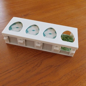以下が含まれることがあります： 白のプラスチック製の卵ホルダーで、5つのスロットがあります。各スロットには卵の形をした切り抜きがあり、切り抜きには緑と白の模様の紙が貼られています。
