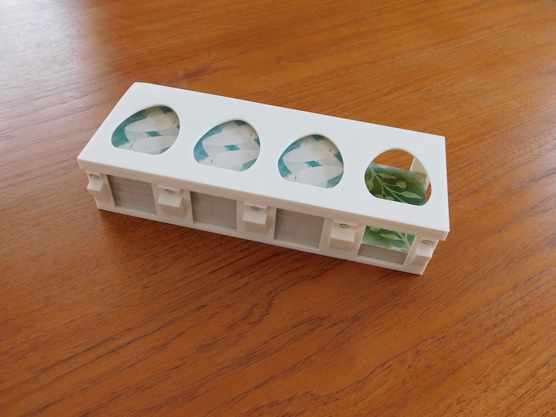 以下が含まれることがあります： 白のプラスチック製の卵ホルダーで、5つのスロットがあります。各スロットには卵の形をした切り抜きがあり、切り抜きには緑と白の模様の紙が貼られています。