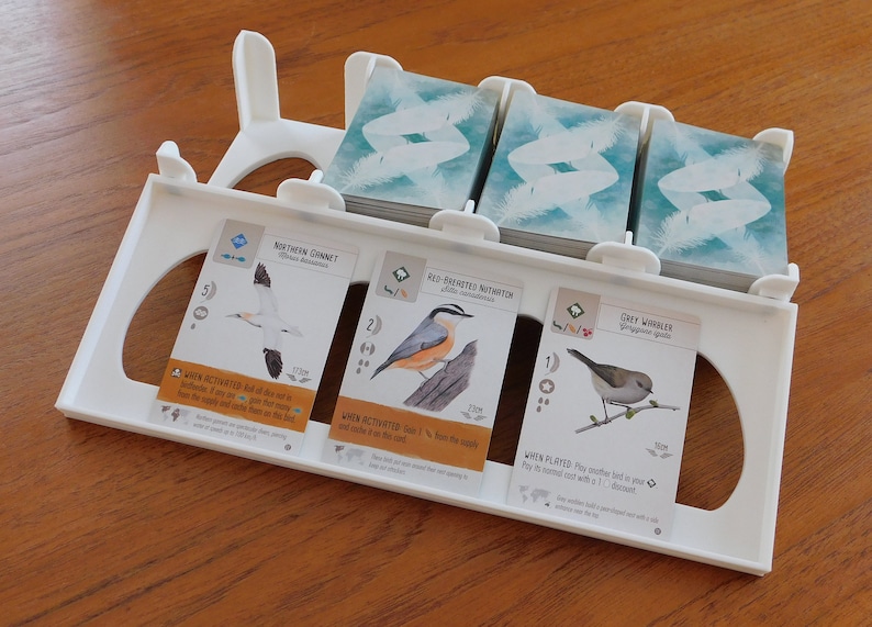 以下が含まれることがあります： 3つのカードスロットが付いた白いプラスチック製のカードホルダー。各スロットには、鳥の絵、名前、特徴の説明が書かれた異なる鳥のカードが入っています。カードは、Wingspanボードゲームからのものです。