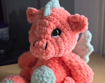 Puede incluir: Un dragón de peluche tejido a crochet en tonos rosa y azul claro. El dragón tiene un pequeño cuerno en la cabeza y grandes ojos redondos.