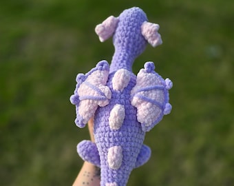 May include: A purple and pink crocheted dragon toy with a long tail and wings. The dragon is held in a person's hand.