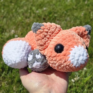 May include: A crocheted fox toy in shades of orange, white, and gray. The plush fox has black eyes and is lying down. The toy is held in a hand against a blurred green background.