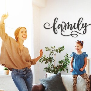May include: A family of three dances in a living room with a wooden sign that reads "family" on the wall. The family is wearing casual clothing and smiling.