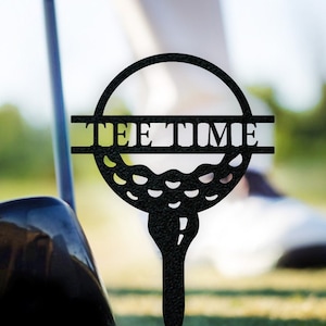 May include: Black metal golf ball tee with the words "Tee Time" cut out in the center. The tee is inserted into a green grassy surface.