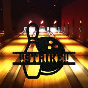May include: Black metal silhouette of a bowling pin and ball with the word "STRIKE" in the middle. The silhouette is set against a blurry background of a bowling alley.