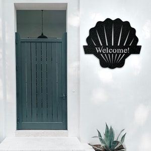 May include: Black metal seashell-shaped welcome sign with the word "Welcome!" on a white background.
