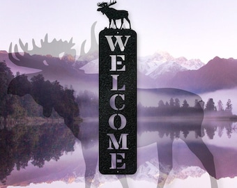 May include: A vertical black metal sign featuring a cutout silhouette of a standing moose at the top. The word WELCOME is vertically laser-cut into the center of the rectangular sign in a bold, serif font with a textured black finish.