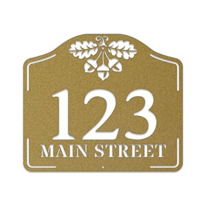 May include: Gold metal house number sign with an oak leaf and acorn design. The sign has the number 123 and the text "MAIN STREET" in white.