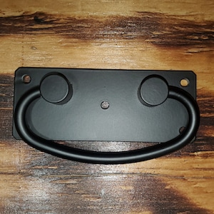 May include: Black metal drawer pull with rounded edges and a D-shaped handle. The pull is mounted on a rectangular plate.