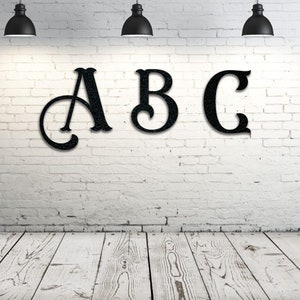 May include: Black metal letters A, B, and C are mounted on a white brick wall. The letters have a decorative, vintage-style font. The wall is illuminated by three black pendant lights, and the floor is made of white wooden planks.