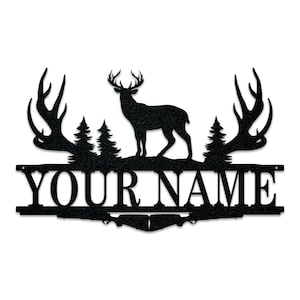 May include: Black metal silhouette of a deer with antlers, standing in front of two pine trees. The deer is facing right. The antlers are curved and point to the left and right. The deer is standing on a black metal base with the text "YOUR NAME" in white letters.