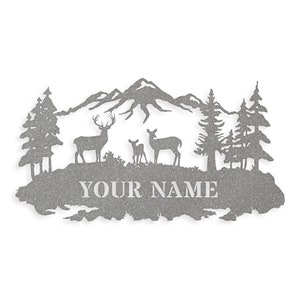 May include: A metal silhouette of a mountain range with three deer, two adult and one fawn, standing in front of a forest. The text "YOUR NAME" is below the deer.