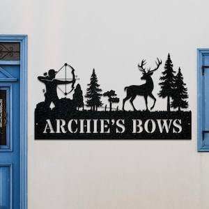 May include: Black metal wall art featuring a silhouette of an archer with a bow and arrow, a deer, and trees. The text "ARCHIE'S BOWS" is displayed below the scene. The artwork is mounted on a white wall.