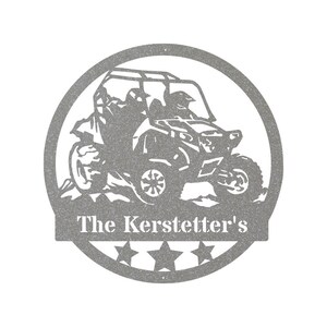 May include: A gray metal silhouette of a side-by-side off-road vehicle with two people inside. The vehicle is driving over a hill and has a white text banner below that reads "The Kerstetter's" with five stars.