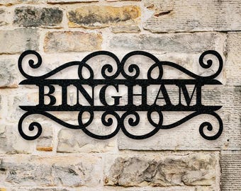 May include: A black metal wall sign featuring the name BINGHAM in capital letters, centered within a horizontal frame decorated with symmetrical scrollwork and swirling flourishes at the top and bottom edges of the rectangular design.
