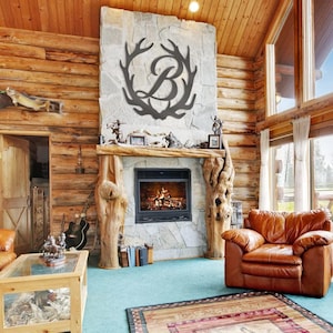 May include: A dark metal wall art piece featuring a large capital letter B centered within a decorative antler frame. The design is mounted on a light-colored stone fireplace chimney breast above a black metal fireplace insert in a room.