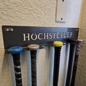 May include: A black metal baseball bat rack with the name "HOCHSTETLER" cut out in white lettering. The rack holds several baseball bats with blue, yellow, and black grips. The rack is mounted on a textured wall.