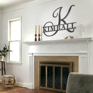 May include: A dark-colored metal wall sign featuring a large, stylized capital letter K with the name KIMBALL integrated into the center. The sign is mounted on a white wall above a fireplace mantel, displaying a clean, cut-out design.