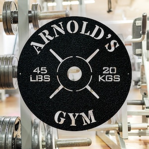 May include: Black metal weight plate with white lettering that reads "Arnold's Gym" and "45 LBS 20 KGS".