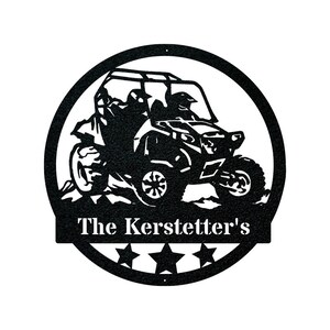 May include: Black metal silhouette of a side-by-side off-road vehicle with two figures inside. The vehicle is driving over a hill. The words "The Kerstetter's" are written below the vehicle in a white banner.