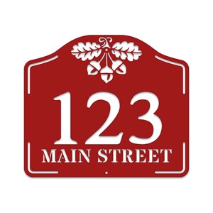 May include: Red metal house number sign with white lettering. The sign features the number 123 and the street name "MAIN STREET" with an acorn and oak leaf design at the top.
