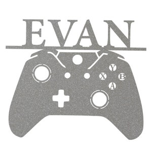 Pode incluir: Um recorte de metal prateado de um controlador de videogame com o nome "EVAN" acima.