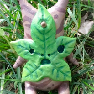 May include: A small, hand-crafted figurine of a character from the video game "The Legend of Zelda: Breath of the Wild". The figurine is made of brown clay and painted with green, brown, and white details. It is wearing a large, green leaf on its head.