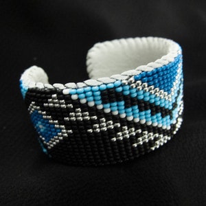 May include: A beaded bracelet with a geometric pattern in black, white, and blue colors. The bracelet is made of small beads and has a white leather backing.