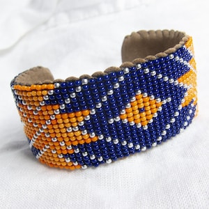 May include: A beaded bracelet with a blue, orange, and silver geometric pattern. The bracelet is made of leather and has a suede lining.