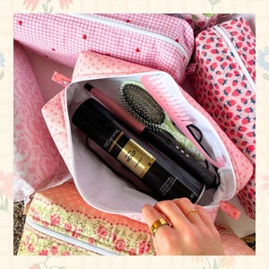 May include: Pink cosmetic bag with a white interior, holding hair care products. The bag is open, revealing a black hairspray can, a brush, and a comb. Other bags with floral and gingham patterns are visible.