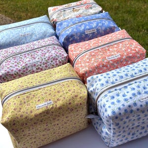 May include: A collection of rectangular fabric pouches with floral patterns in various colors, including blue, pink, yellow, and peach. Each pouch has a white zipper and a small label that reads "Cute and the Spudles".