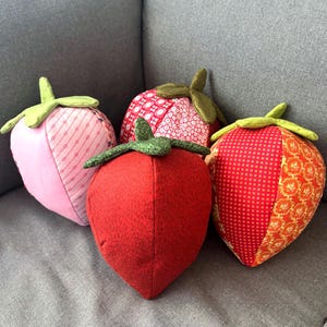 May include: Four fabric strawberry-shaped decorative items. The strawberries are red, pink, and patterned with green fabric stems. The strawberries are sitting on a gray surface.