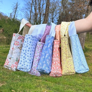 Könnte beinhalten: Eine Sammlung von acht kleinen, gesteppten Stoff-Tragetaschen mit verschiedenen Blumen- und Mustermotiven. Die Taschen sind in den Farbtönen Rosa, Blau, Gelb und Pfirsich gehalten und haben weiße Henkel. Die Taschen werden vor dem Hintergrund von grünem Gras und Bäumen gehalten.