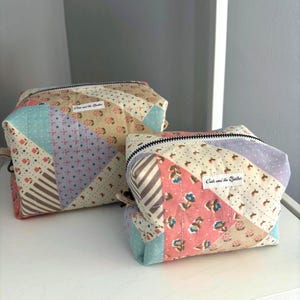 May include: Two patchwork fabric zippered pouches in pastel colors. The larger pouch has a light beige background with pink, blue, and purple accents. The smaller pouch has a similar design with pink floral patterns. Both have black zippers and the text "Cats and the Quilter" on a white label.