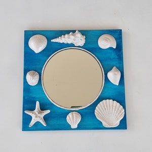 A blue square wooden mirror decorated with white seashells. The round mirror glass is surrounded by white seashell sculptures of various shapes and sizes.