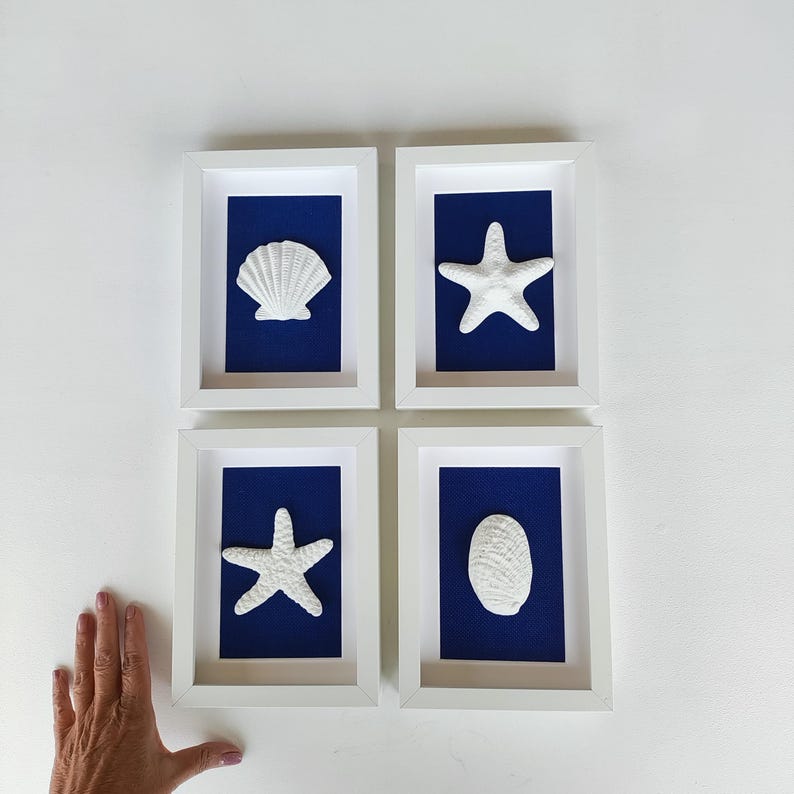 Cuatro obras de arte costero enmarcadas en blanco con monturas azules. Cada imagen presenta una concha o una estrella de mar blanca sobre un fondo de tela arpillera azul marino.