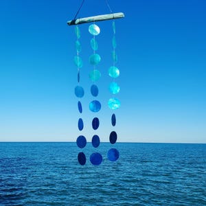Peut inclure: Un carillon éolien suspendu, composé de coquillages ovales bleus irisés, suspendus à une poutre en bois naturel. Les coquillages présentent différentes nuances de bleu. Le fond montre un ciel bleu clair et la mer.