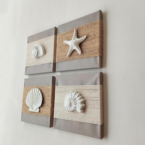 Op de afbeelding: Een set van vier vierkante wandpanelen met een gladde grijze afwerking. Elk paneel heeft een horizontale band van gewikkeld touw in beige of crèmekleurige tinten. In het midden van elke touwband is een witte, driedimensionale schelp of zeester bevestigd.